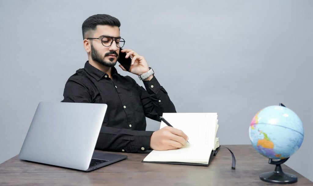 Man on phone writing at desk with globe.